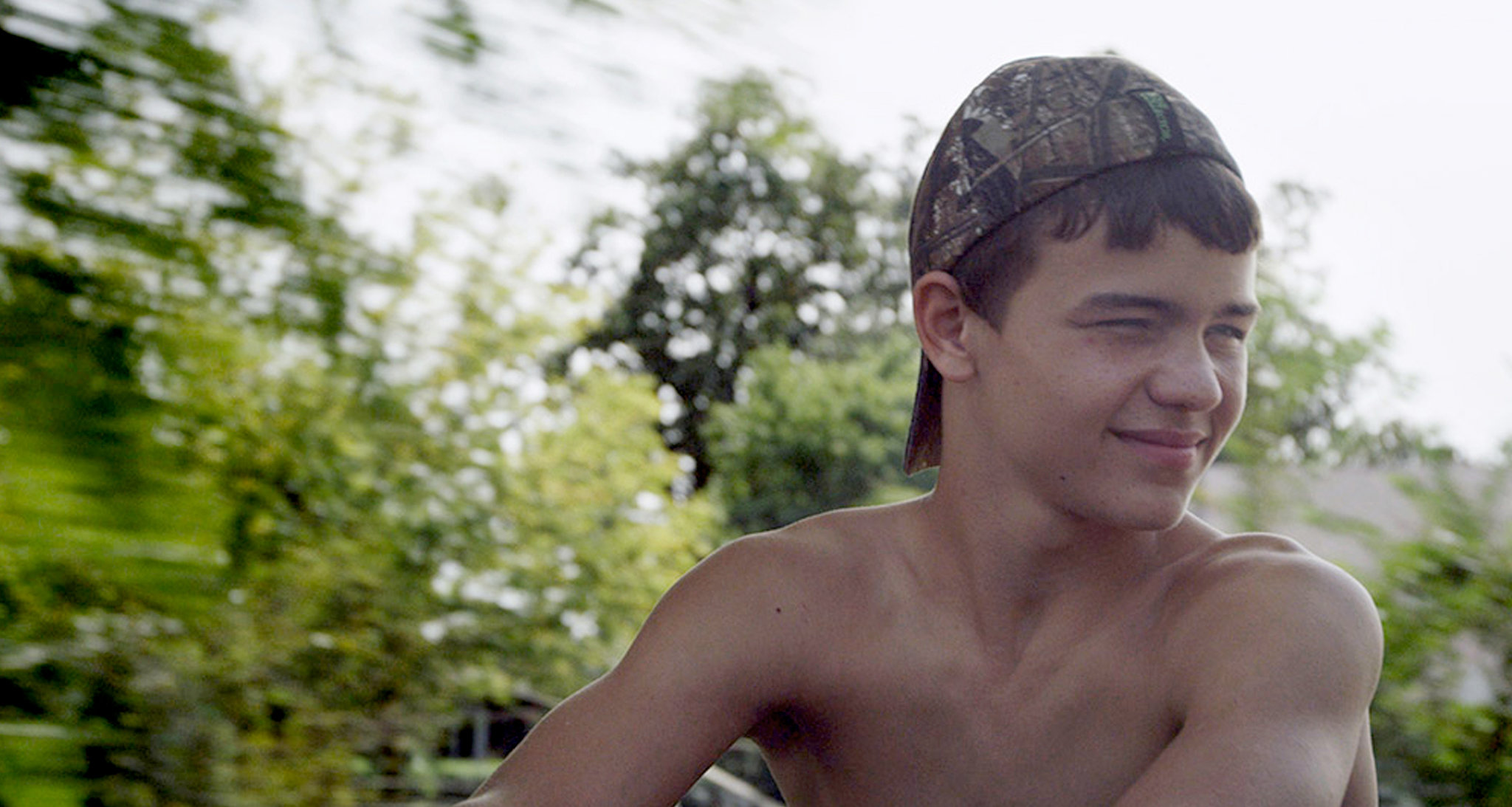 shirtless man looks to the left and smiles around plants