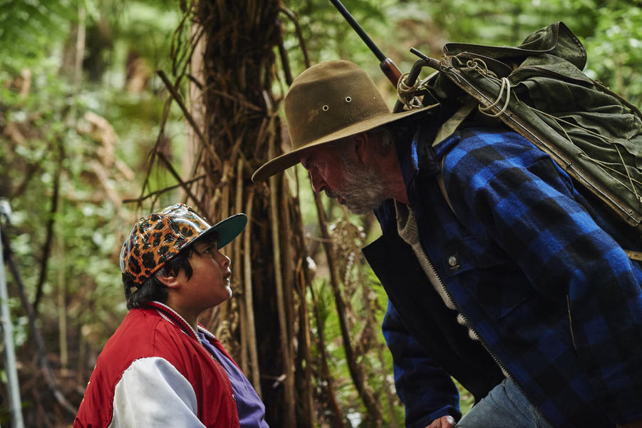 Gray-bearded man with hat and backpack leans down to talk to boy in red jacket and cap.