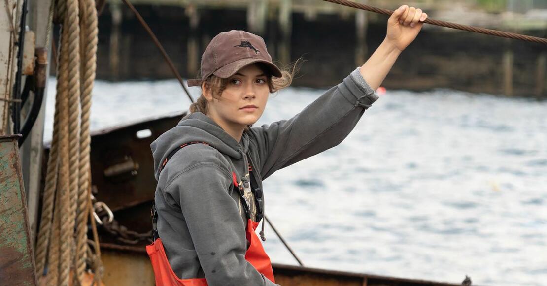 Young white woman in cap, gray hoodie and red protective trousers holds onto a rope while apparently sitting in a boat