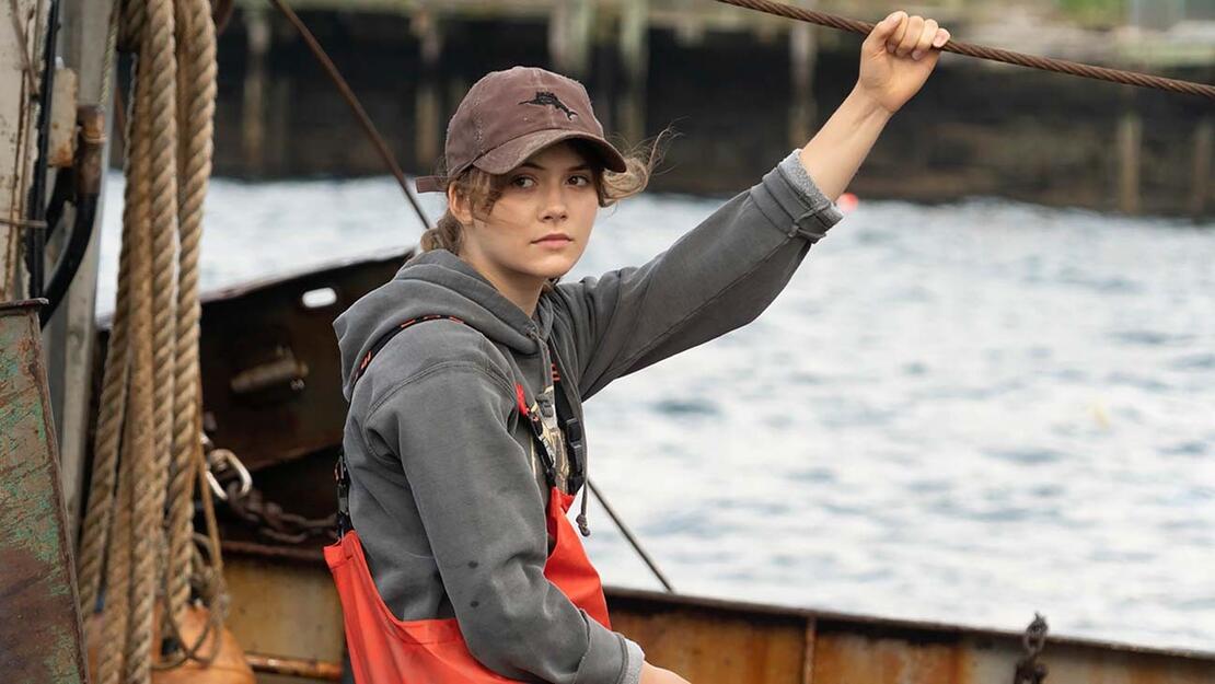 Young white woman in cap, hoodie, and waterproof pants hangs on to a rope on a boat