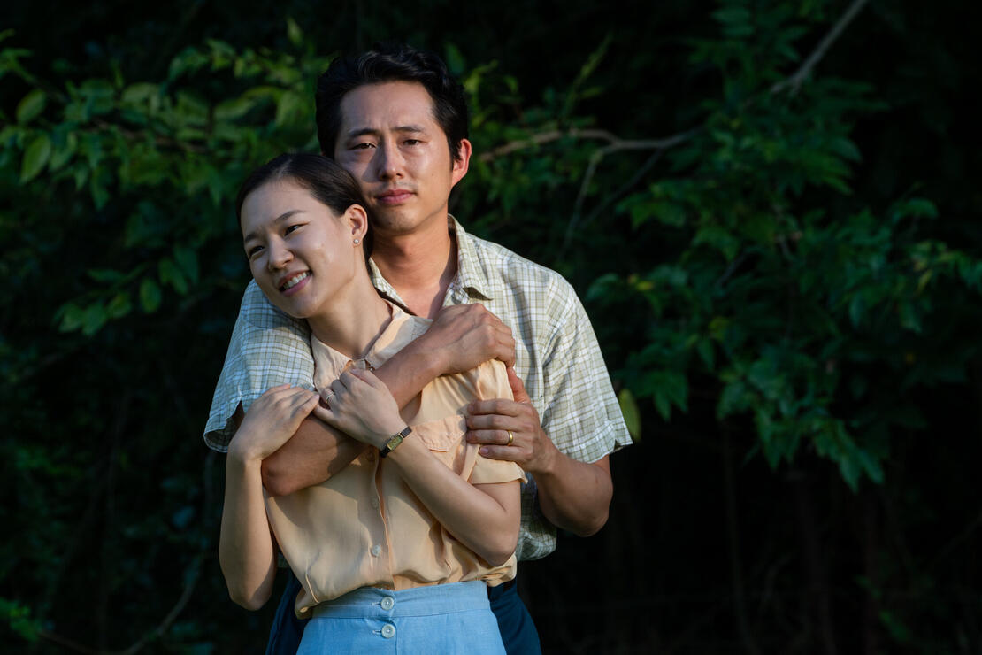 Asian couple embraces, both facing the camera, in front of a tree.