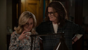 A blonde women (Elizabeth Banks) sips coffee from a white mug next to a brown-haired woman (Sigourney Weaver) with glassed and a white top with a black button down shirt over.