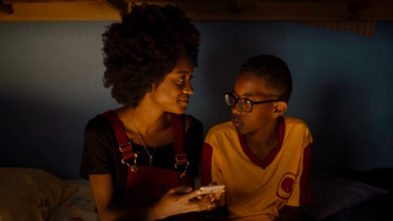 Black woman in overalls and afro smiles at the young Black boy to her left, a bespectacled boy who stares solemnly back.