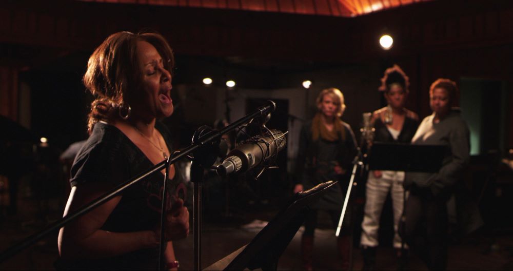 Woman, in left foreground, sings into an elaborate microphone setup, while three other women stand in the background.