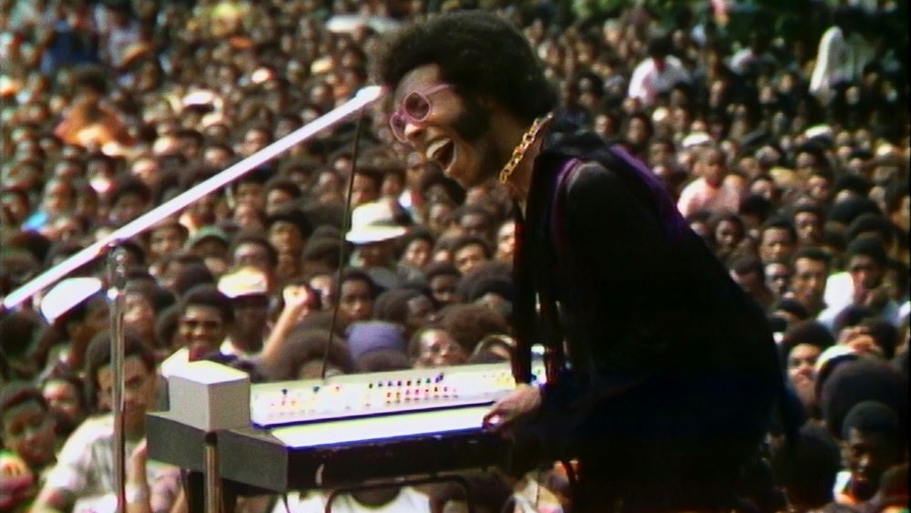 Gleeful Black man, with gold necklace and pink eyeglasses, plays a keyboard or sound board in front of a huge audience outdoors.