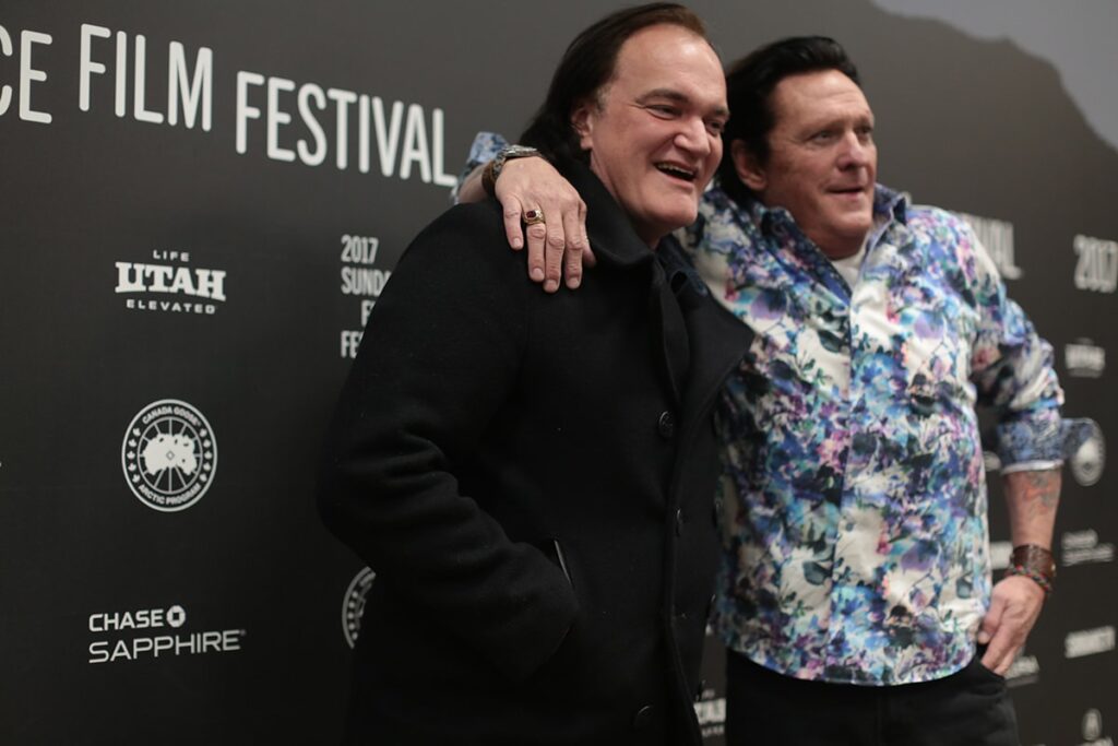 Two smiling men stand in front of a film festival backdrop. The man on the right, in a flowered shirt, drapes his arm over the shoulders of the man on the left, who is dressed in all black.