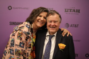 A tall, dark-haired woman smiles, with her arm around a man's shoulders, left hand placed on his shoulder. The man, dressed in a dark suit and tie, a yellow rose on is jacket pocket, also smiles. Their heads are touching.