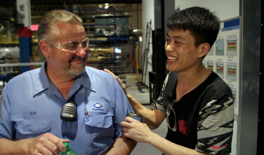 A bearded white man in a blue uniform shirt and safety glasses and an Asian man in a black Nike shirt smile at each other, in an apparent factory setting.