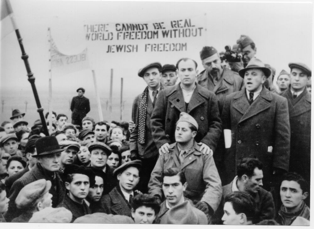 A crowd of men in 1940s-style coats and clothing stand in front of a banner that says "There Cannot Be Real World Freedom Without Jewish Freedom."