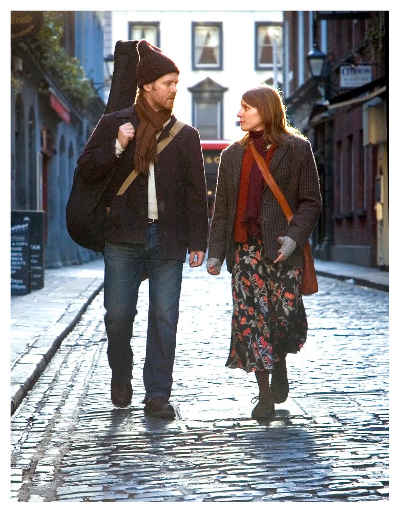 Man with guitar on his back and woman walk down cobblestone city stree.