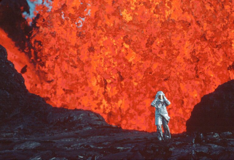 Small person in flame retardant suit in front of bright orange volcano lava blast