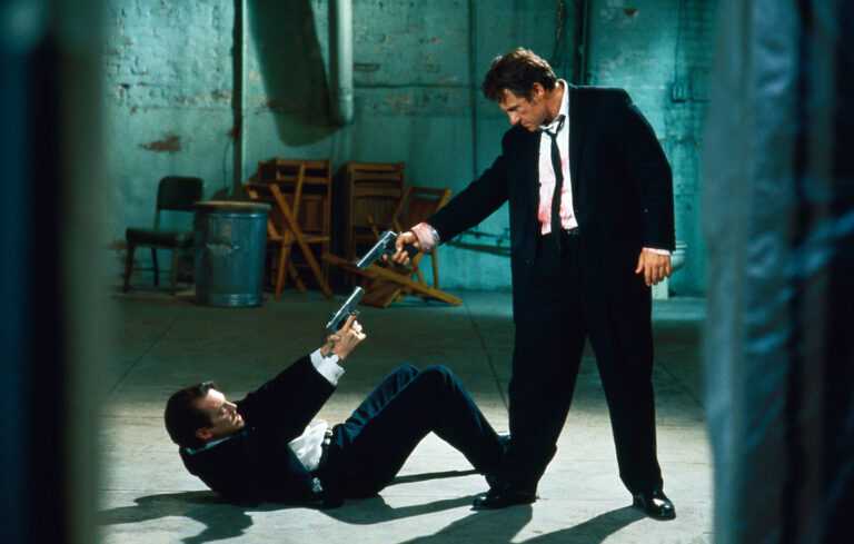 A man in a suit, with what appears to be blood on his shirt, points a semi-automatic pistol and a man lying on his back on the floor, who points a semi-automatic pistol at the standing man. They appear to be inside a warehouse type of building.
