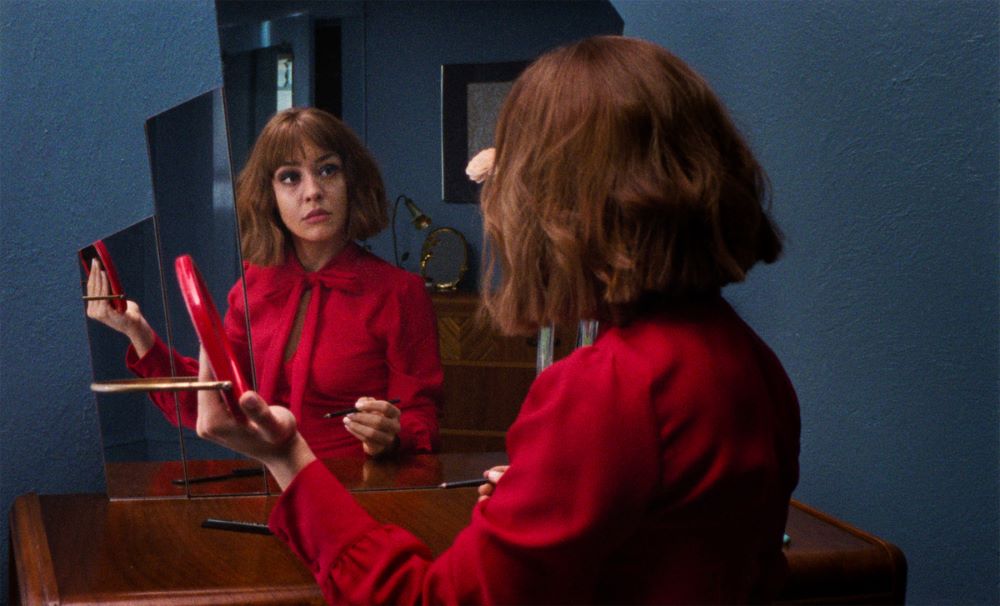 A young woman in a red dress looks into a series of mirrors.