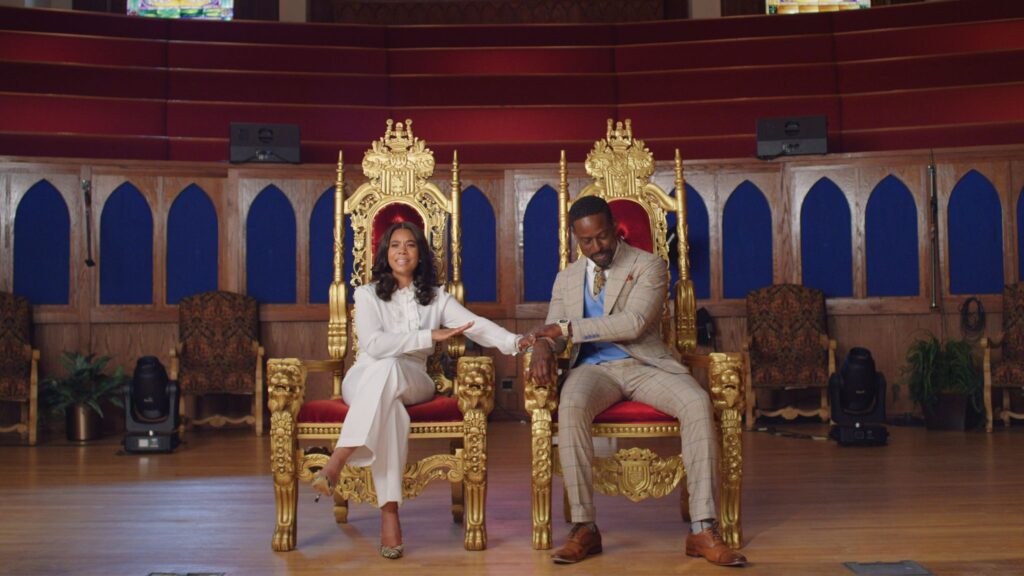 A dressed-up Black couple sit in side-by-side gold and red velvet thrones