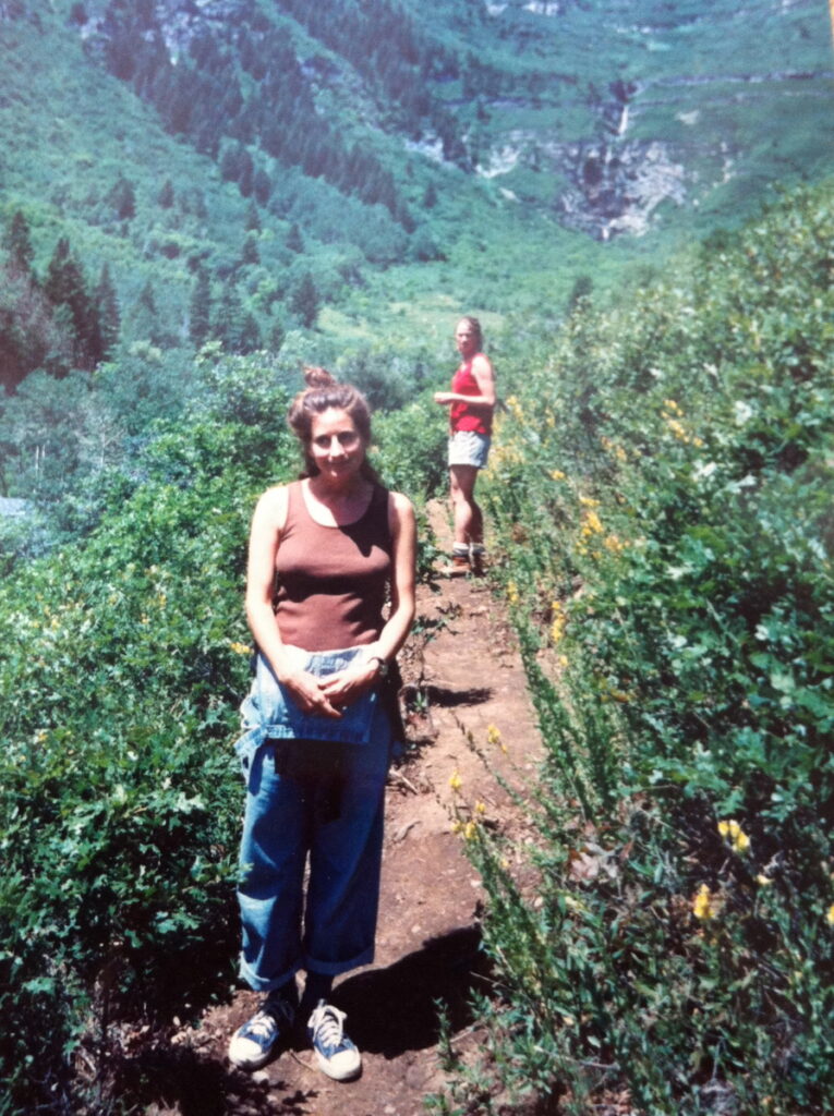 A woman stands in the foreground on a trail in mountainous terrain.