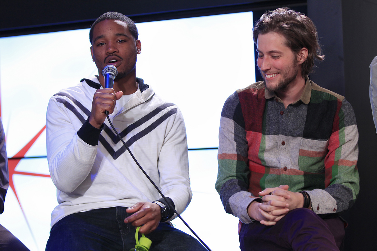 Two men sitting in front of a screen or background, one holding a microphone, the other smiling