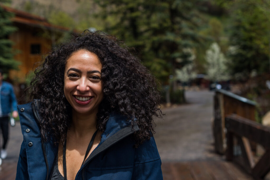 A woman with curly black hair stands in front of trees in an outdoor setting.