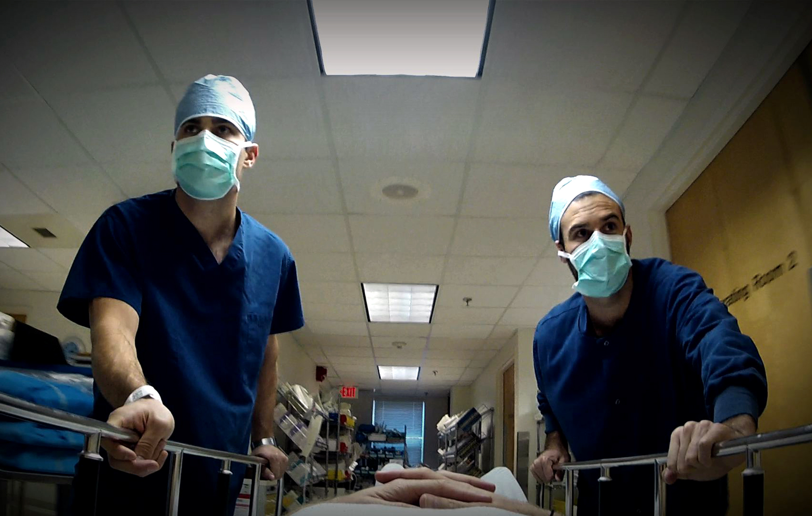 Two ER medical professionals in blue with masks are rushing through the emergency room with a gurney.