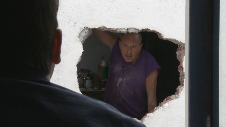 Man peers from a hole in the wall at another man
