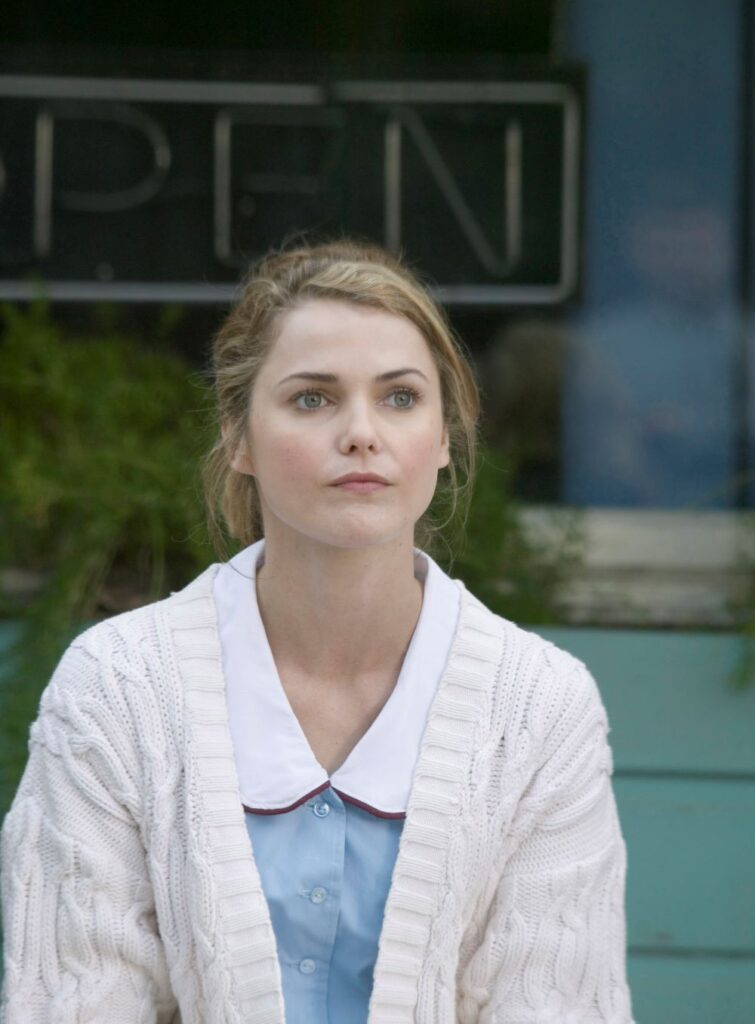 A woman in a waitress uniform, with a sweater worn over it, is in front of a business with the "open" sign turned off.