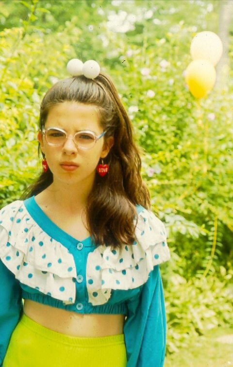 Young girl wearing glasses and a colorful outfit stands outdoors.