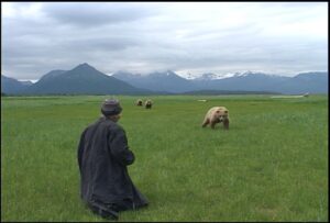 A man is kneeling on the grass as a white grizzly bear approaches him