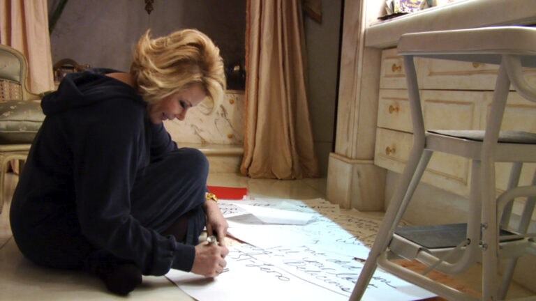 A blonde woman dressed in black is kneeling on the floor looking at pages of notes spread out.