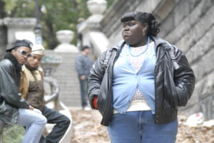 A young black girl with short bangs wearing a baby blue sweater and a black coat is walking with her hands in her pockets as two bystanders look on.