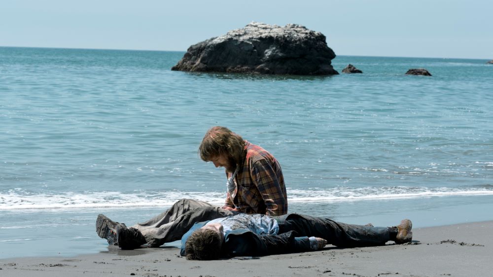 Bearded man sits on shore next to what appears to be a dead body.