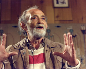 Gray-haired and bearded man in red and white striped sweater and brown jacket gesturing as he talks