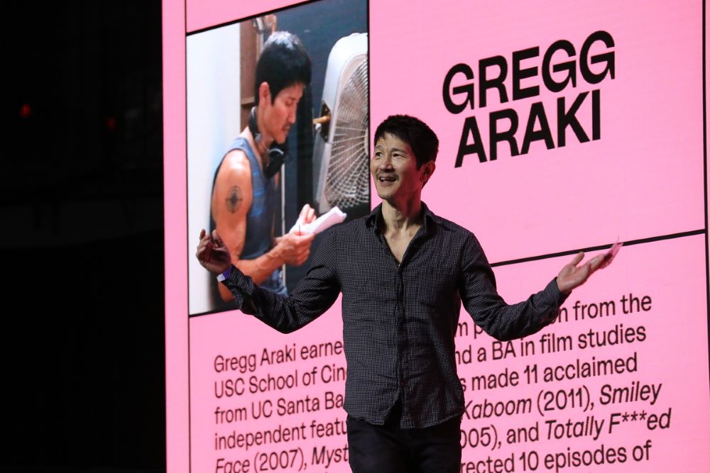 Asian man stands in front of pink board with "Gregg Araki" written on it, as well as biographical information.
