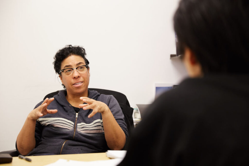 Woman of color sitting behind a desk, gesturing while speaking to a person with back to the camera