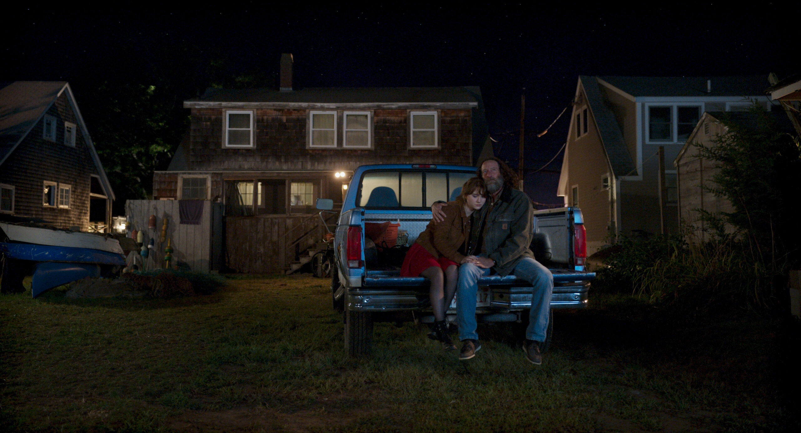 A young woman and older man sit on the tailgate of a pickup truck parked behind a house