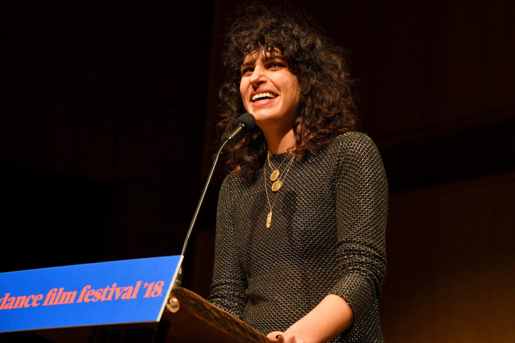 Woman with dark, curly hair standing behind a 2018 Sundance Film Festival podium