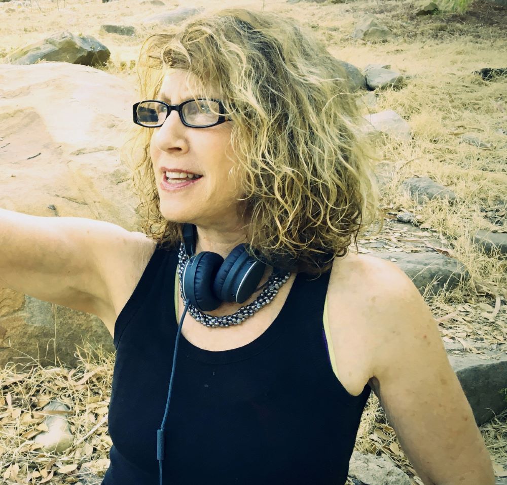 Blond woman in eyeglasses, and with headphones hanging around her neck , gestures