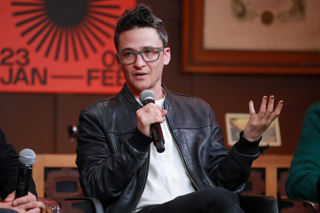 Person with pompadour in glasses and leather jacket gestures while speaking into a microphone