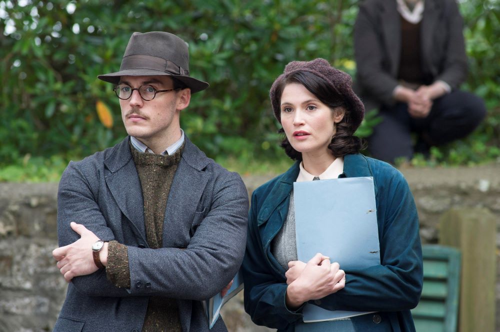 Man in hat and woman holding a sheaf of papers watch something intently.