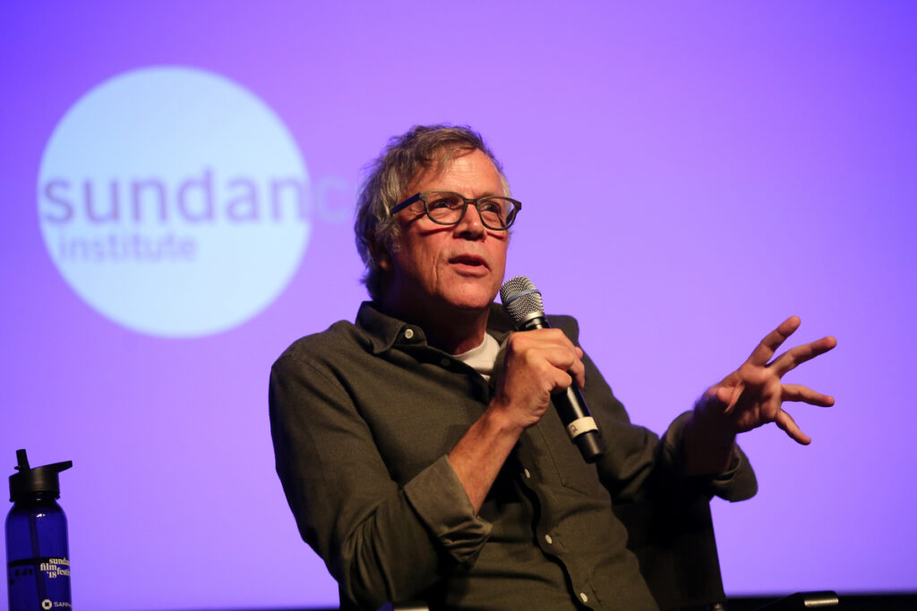 Man with gray hair and glasses gesturing while speaking into a microphone