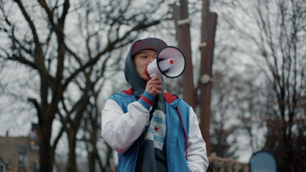 Asian man in hoodie and cap speaking through a megaphone in an outdoor setting