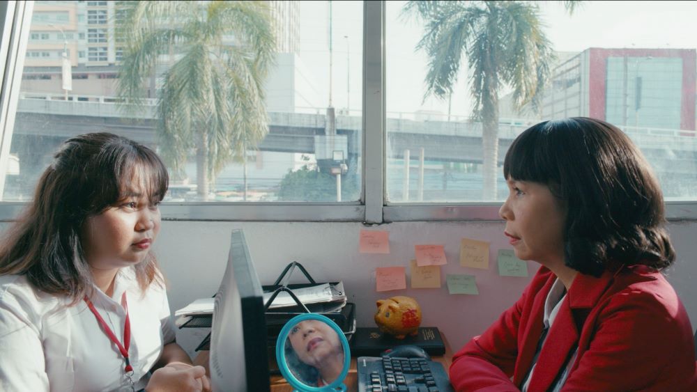 Two women sit opposite each other in an office setting, with palm trees outside the window