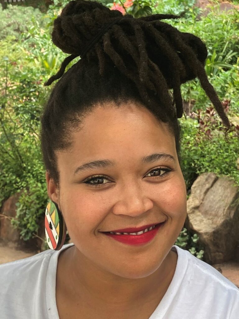 Headshot of woman of color with bright smile, large earrings, and dreadlocks piled on top of head