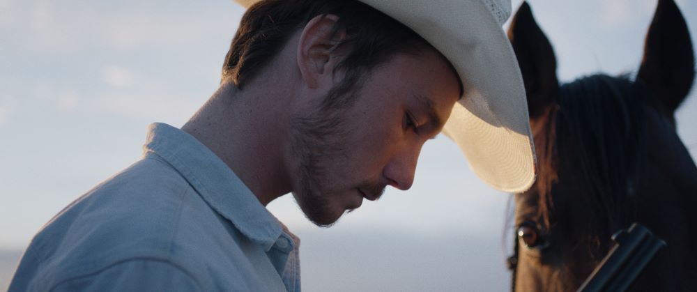 Young bearded man in light cowboy hat looks down pensively, the eye and head of a horse to the rightt