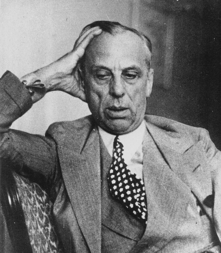 Man in suit and tie, eyes downcast and body leaning slightly to his right, hand placed on his head.