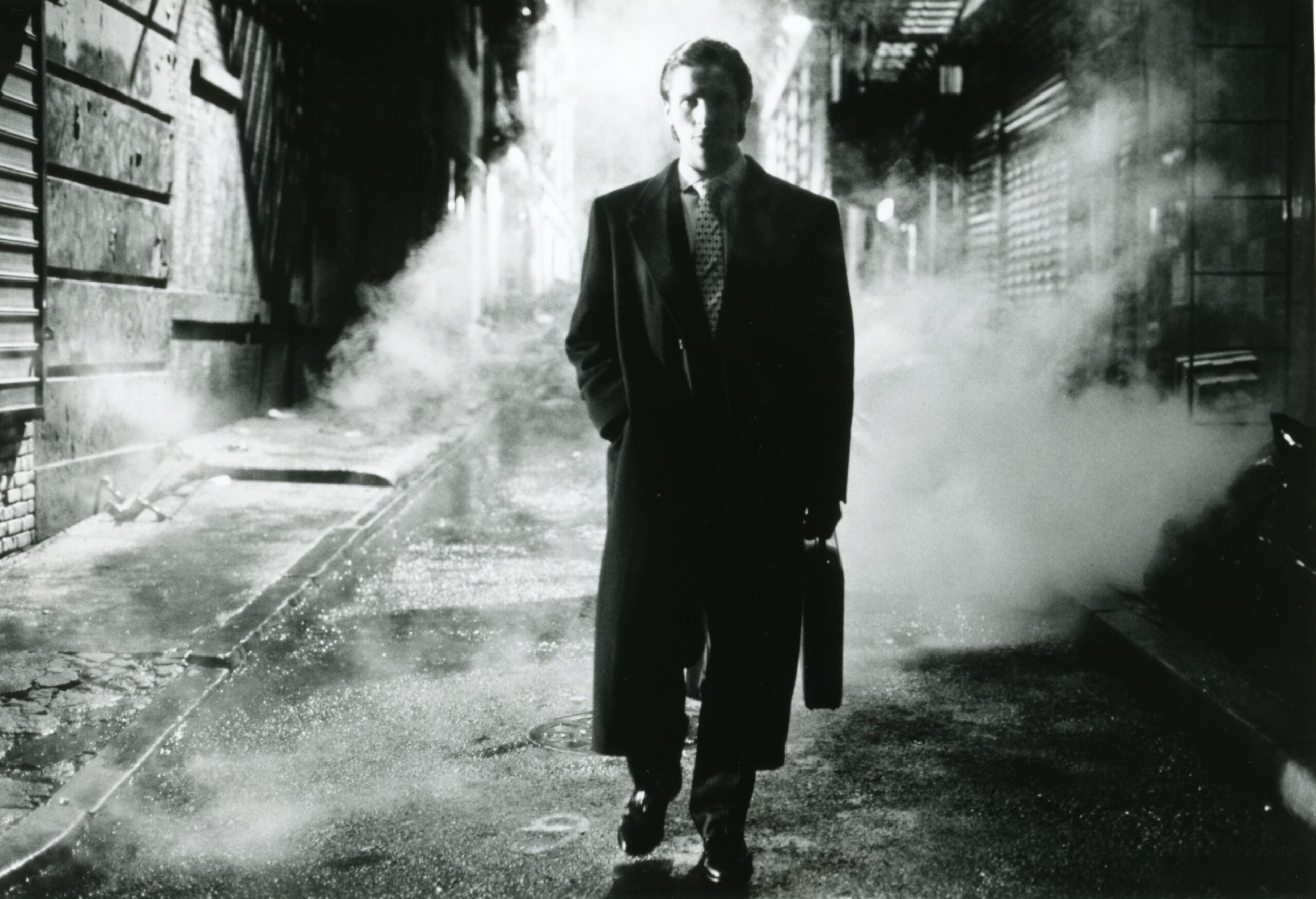 A black and white still of a man (Christian Bale) in a black suit with a long black coat, holding a briefcase. He is walking down a dark alley as smoke and fog surround him.