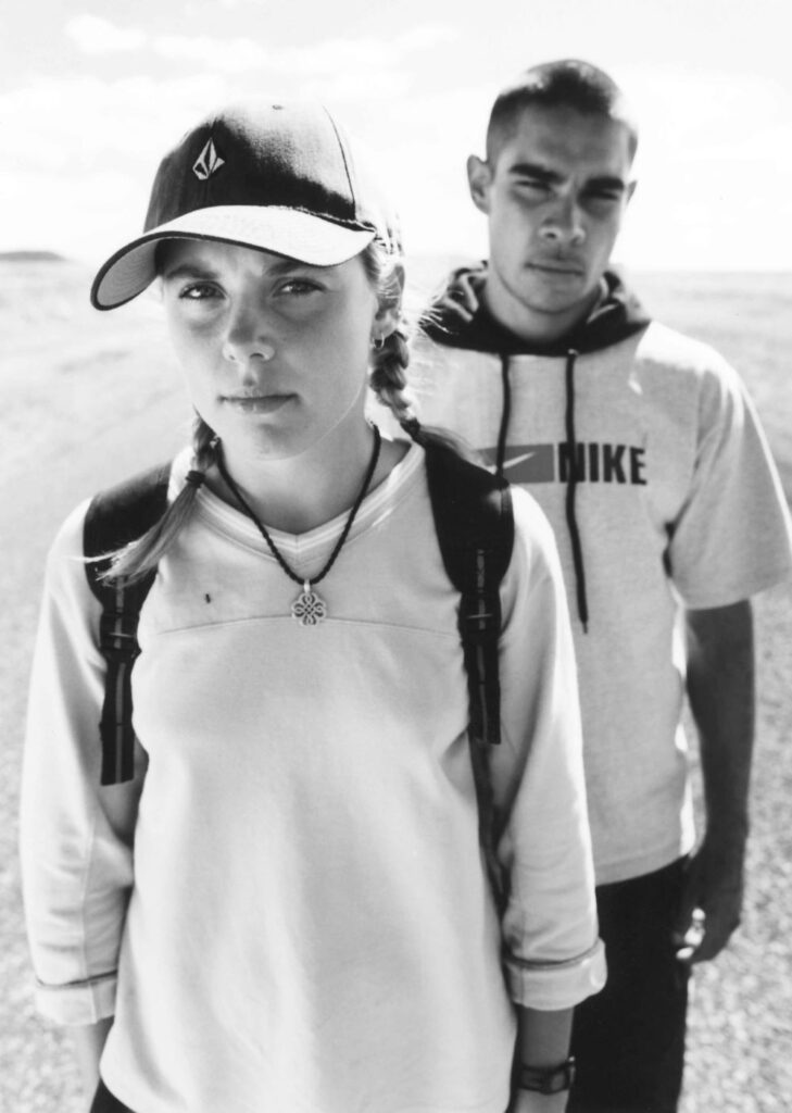 A woman in a cap and backpack stands in front and to the side of a man in a short haircut and short-sleeved hoodie, in what appears to be an arid landscape