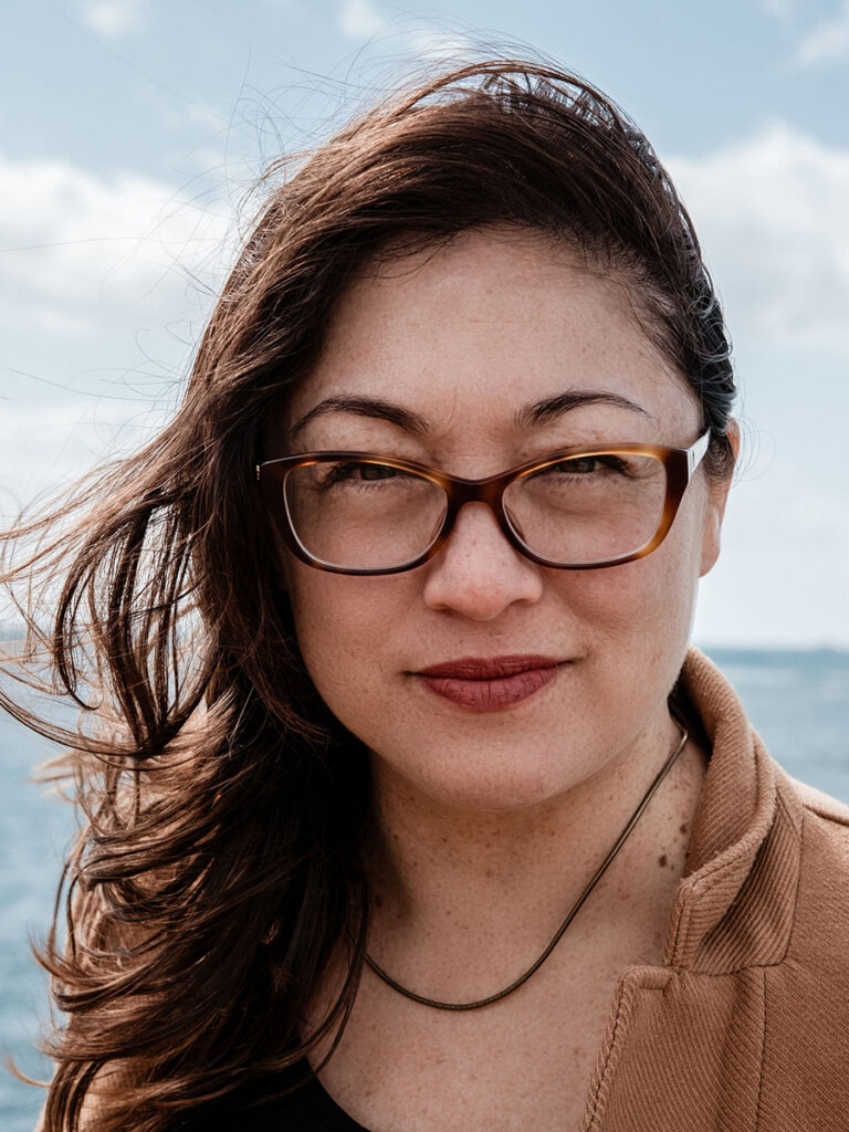 Asian women in eyeglasses and wind-blown long, brown hair, with what appears to be an ocean in the background