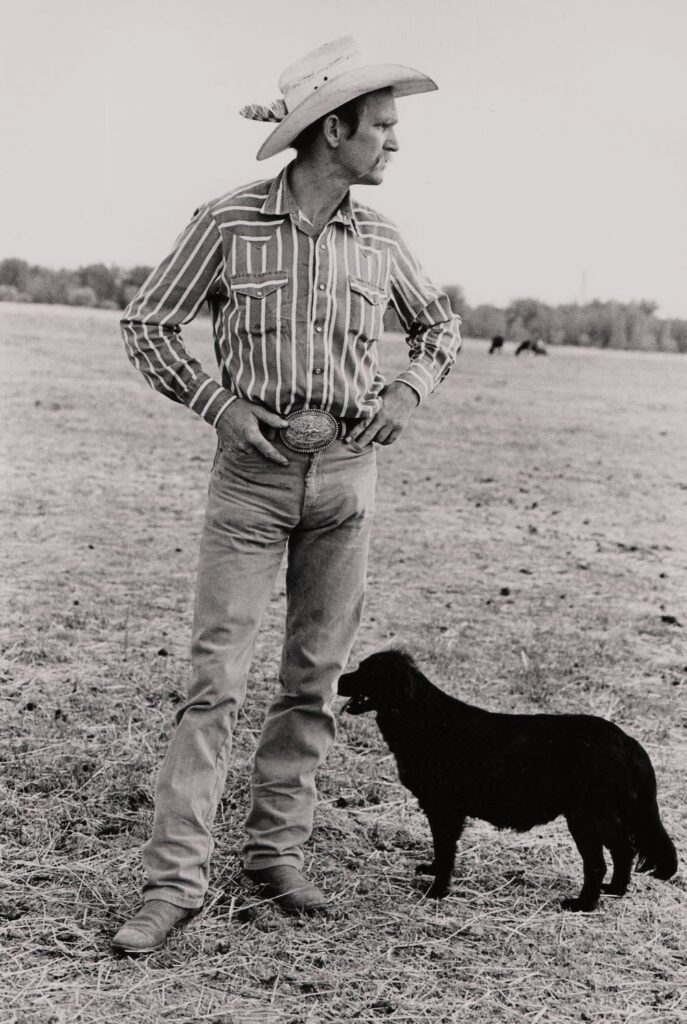 Man in light cowboy hat with feature, a striped western shirt, jeans, and boots stands in the dirt and dry grass, with a medium-sized dark dog at his side.