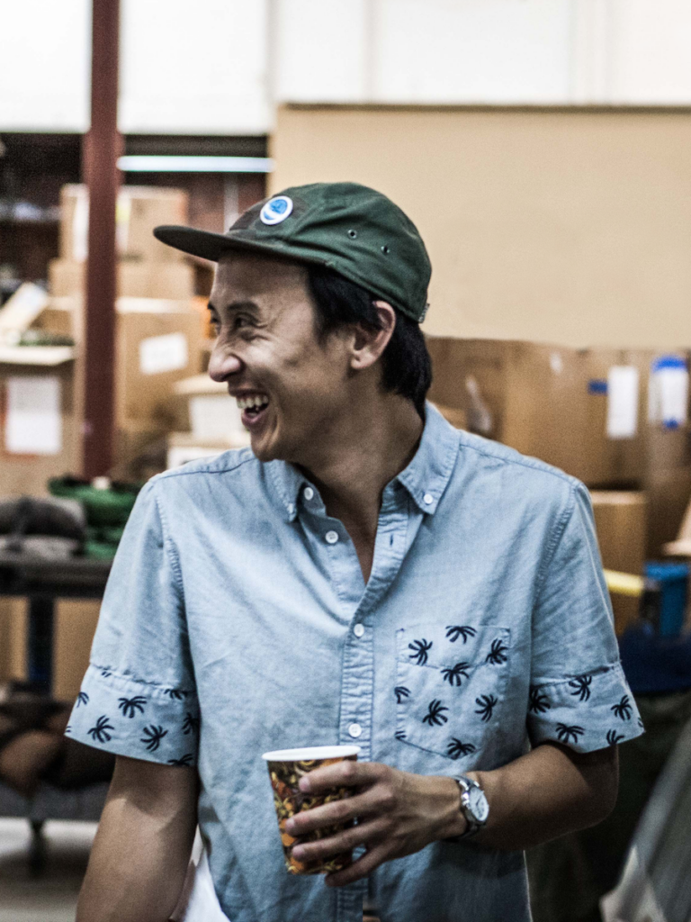 Man in green cap, light blue shirt, holding a cup, looking to side and laughing