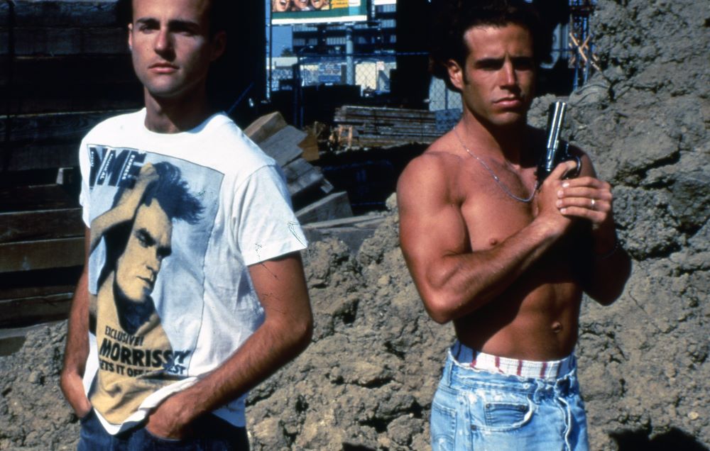 A man in a white Morrissey T-shirt and a shirtless man holding a revolver stand side by side