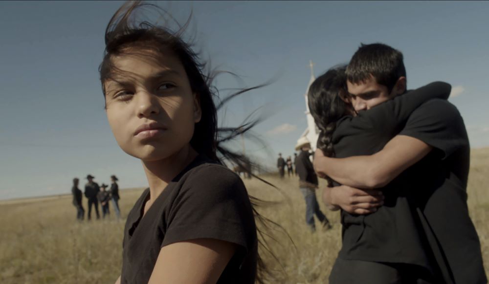 Young native girl stands in a prairie, with a young man and woman embracing in the near background, and mother people in westernwear and a church in the background